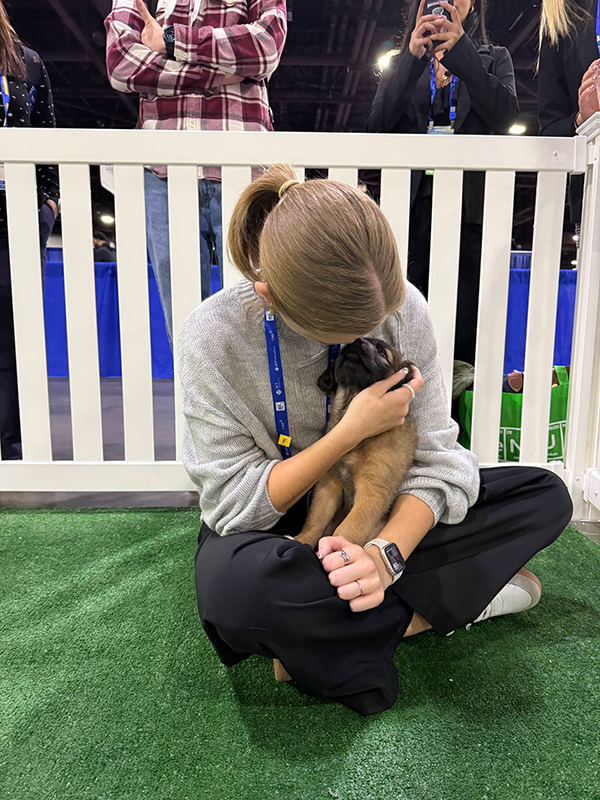 A student sitting down at the ACS conference, smiling and cuddling a small brown puppy.